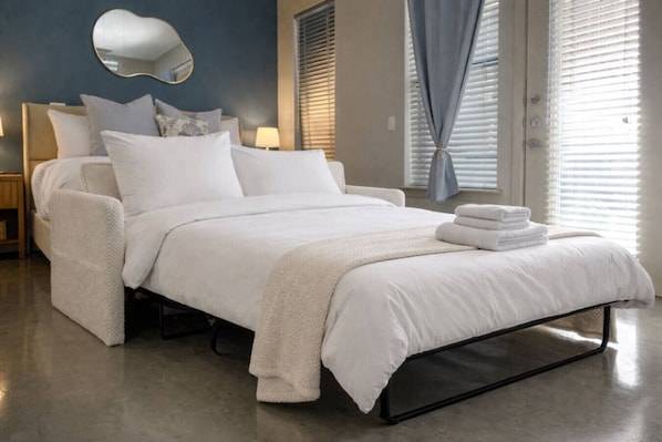Un dormitorio moderno con cama, almohadas, una mesita de noche, una lámpara y una ventana con persianas.