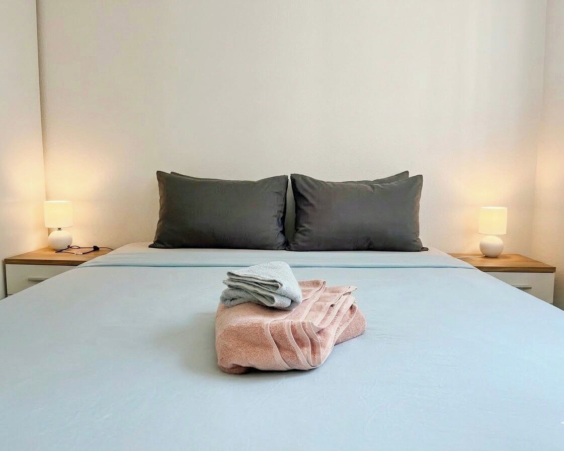 A hotel room with a neatly made bed, two bedside lamps, and a folded towel on the bed.