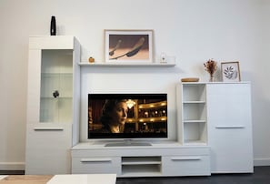 A modern living room with a flat-screen TV mounted on a white entertainment unit, a glass cabinet, and a small plant.
