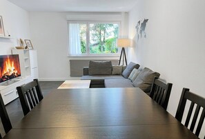 Moderni olohuone, jossa on takka, sohva, ruokapöytä ja ikkuna, josta avautuu näkymä vehreään maisemaan.