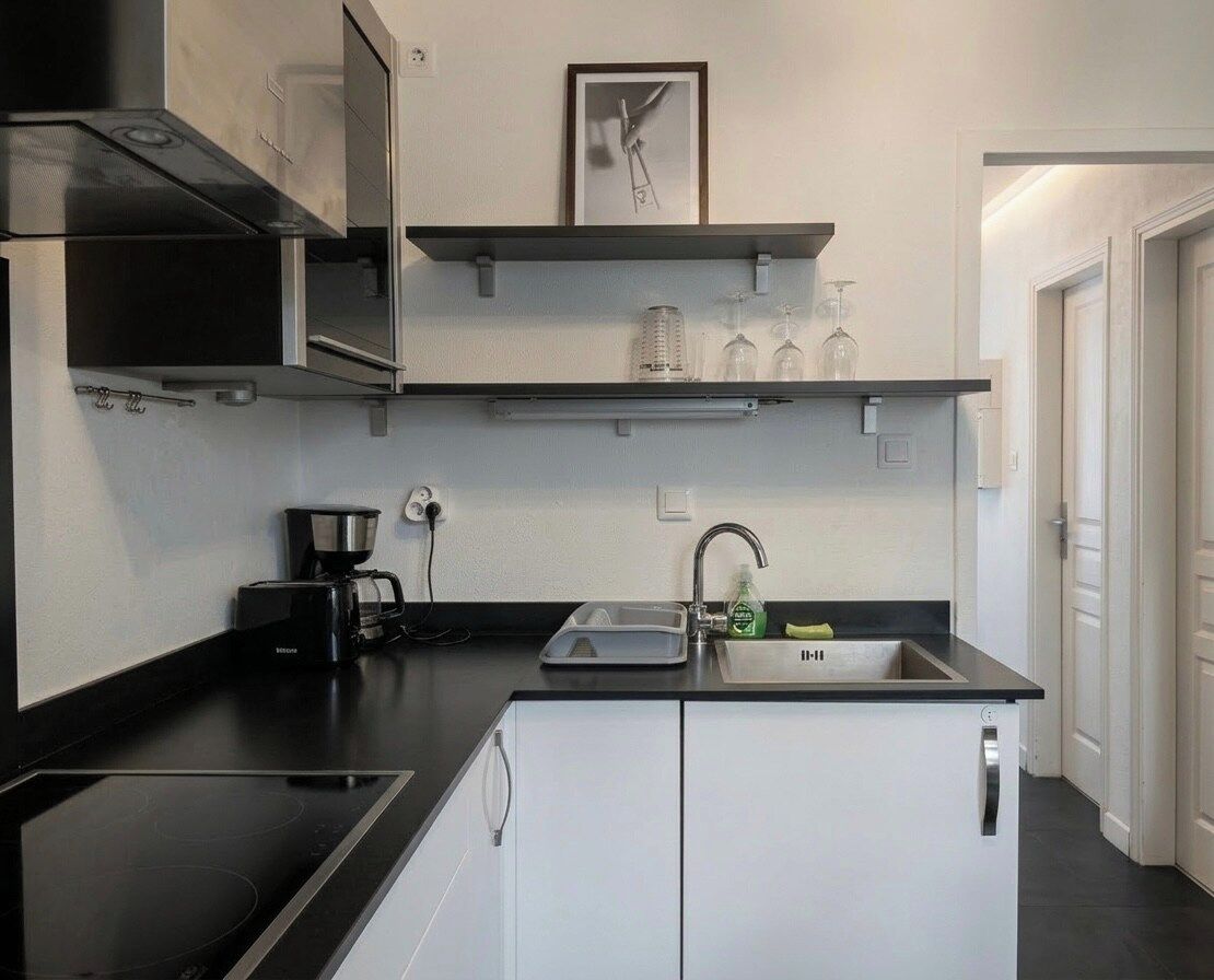 Une cuisine compacte dotée d’un plan de travail noir, de meubles blancs, d’une cuisinière encastrée, d’une machine à café et d’un évier avec un robinet.