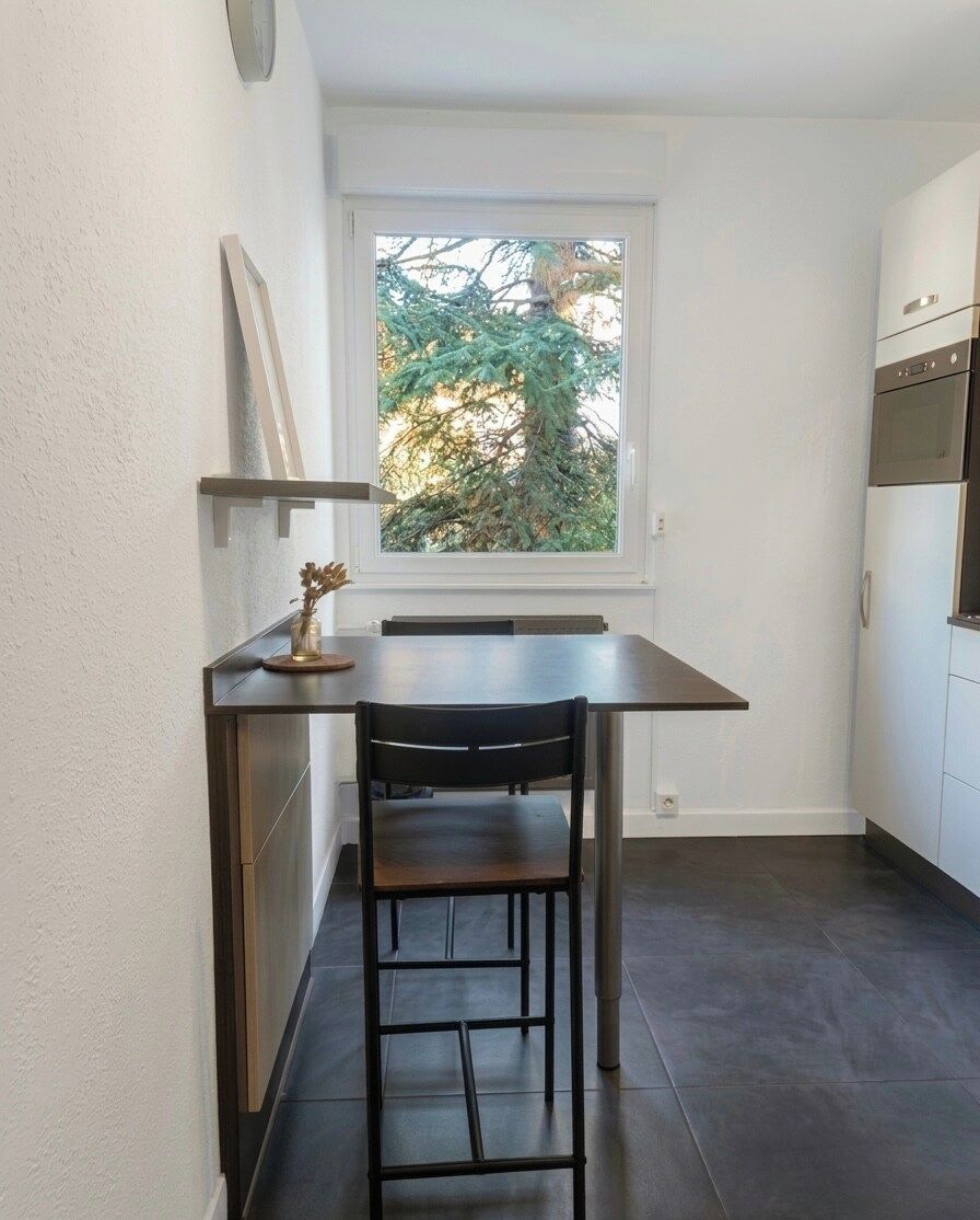 Een compacte keuken met een eettafel en stoelen, een raam met uitzicht op bomen, en inbouwapparatuur.