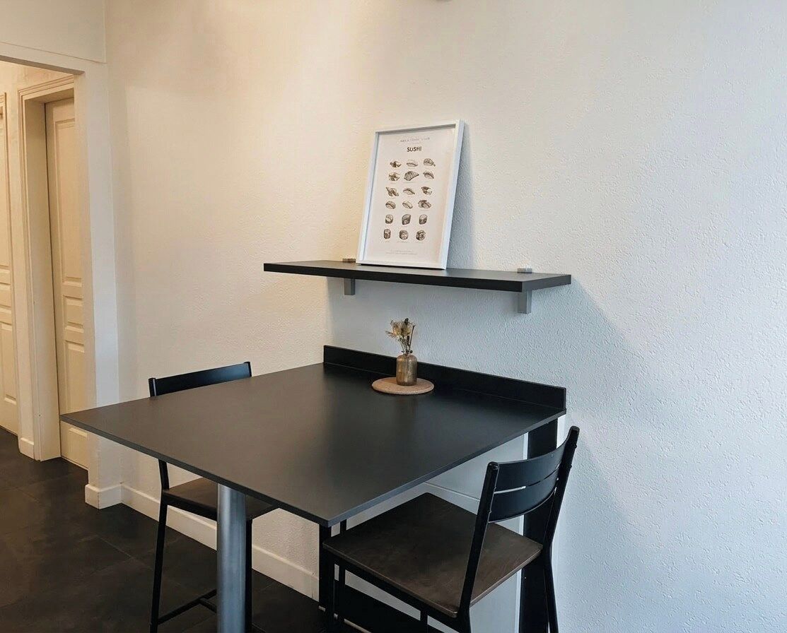 Un coin repas avec une table et des chaises noires, un menu encadré posé sur une étagère fixée au mur, et un vase avec des fleurs sur la table.