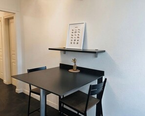 Ruokailutila, jossa on musta pöytä ja tuolit, seinälle kiinnitetty hylly, jonka päällä on kehystetty ruokalista, sekä maljakko kukkineen pöydällä.