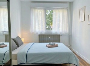 Hotellihuone, jossa on sänky, ikkuna verhoilla, patteri ja peili.