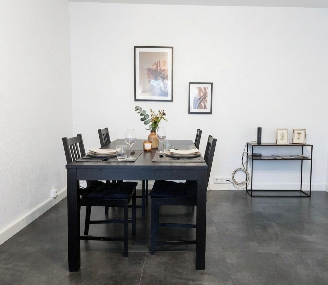 Un coin repas avec une table en bois foncé, dressée pour quatre personnes, entourée de chaises. On y trouve une petite table d’appoint avec un vase et une bougie.