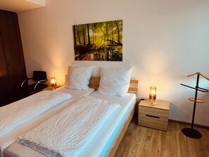 A double bed with white linens, two bedside tables with lamps, a wooden nightstand, and a painting of a forest on the wall. - WeserWaldGlück Vacation Rental - "A Little Getaway" - NEW in Wesertal for 4-5 people (Wesertal)