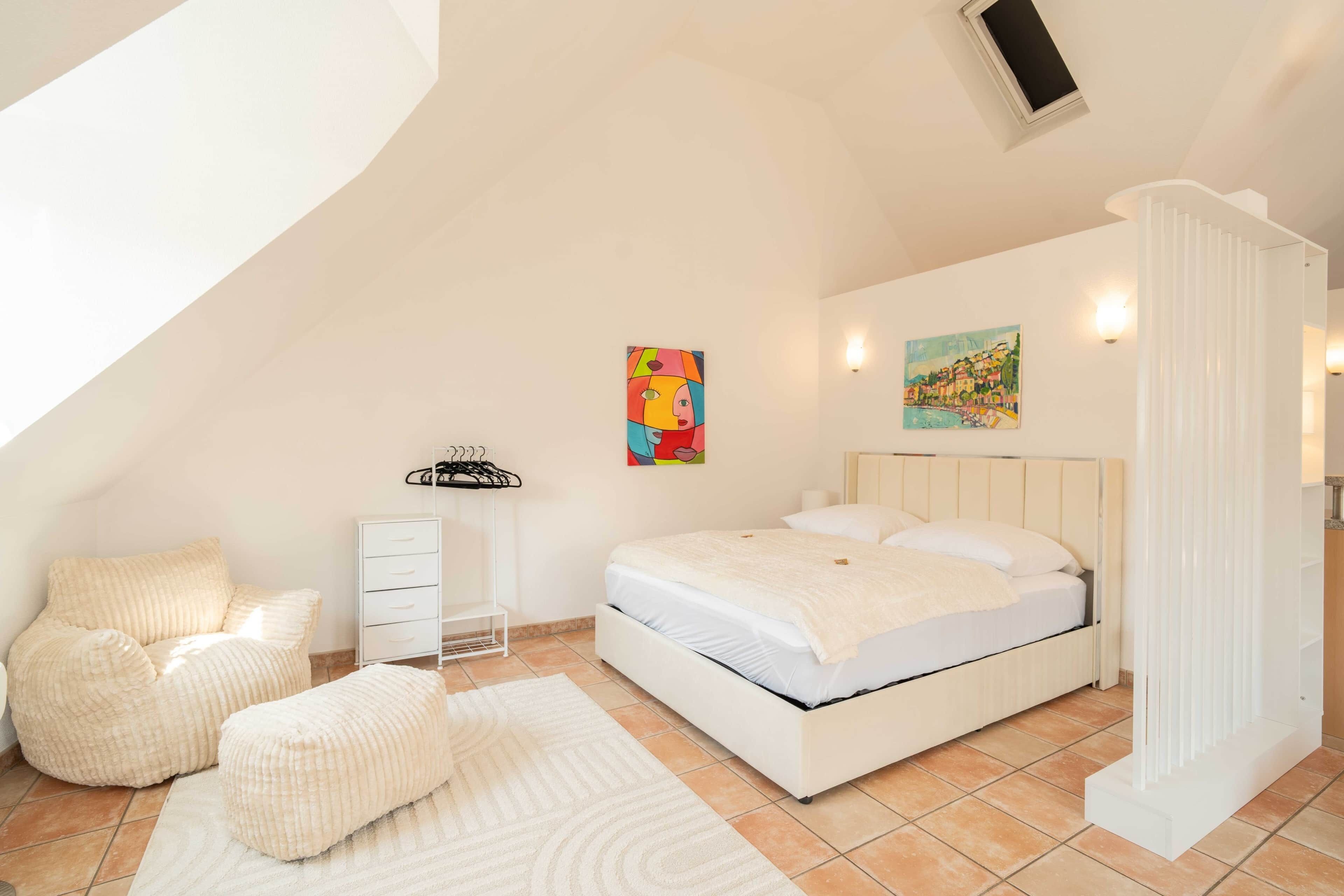 Un dormitorio con una cama, una silla, un aparador y un cuadro en la pared.