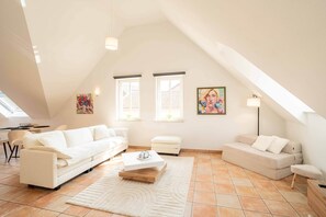 Un vaste espace de vie aménagé dans un style loft, avec un plafond haut, deux fenêtres équipées de stores, un canapé blanc, un canapé plus petit, une table basse et une lampe.