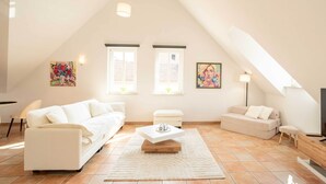 A spacious loft with a high ceiling, white walls, and a sloped roof. There are two white sofas, a small white table, and a floor lamp. - Stylish Apartment | WiFi, Sky TV & Nespresso (Sobernheim)