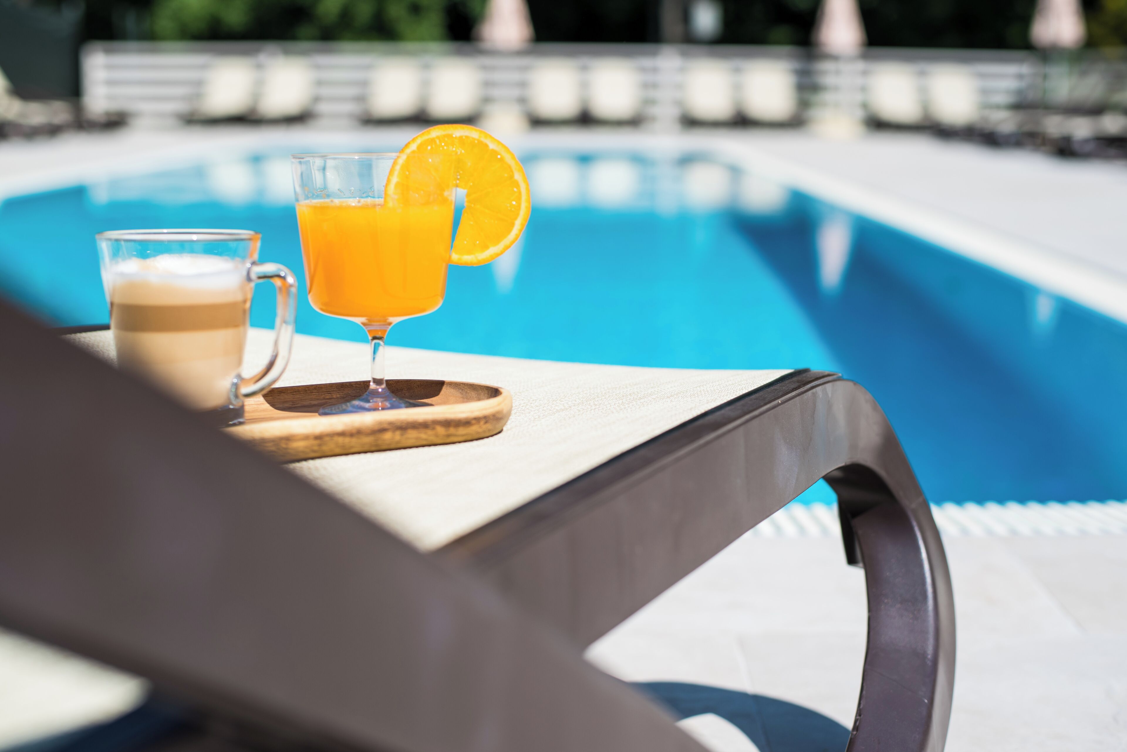 Une scène de bord de piscine, avec un verre de jus d’orange et une tasse de café sur un plateau en bois.