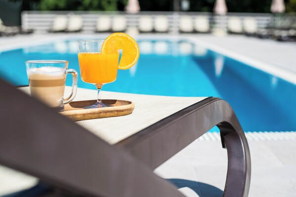 A poolside setting with a glass of orange juice and a cup of coffee on a wooden tray.