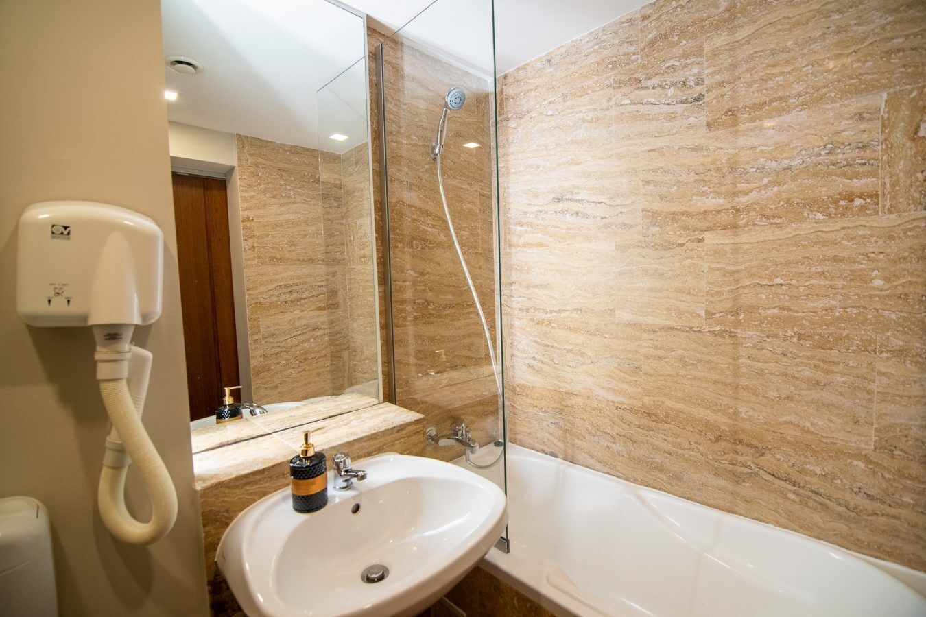 A bathroom with a marble wall, a sink, a mirror, a handheld showerhead, and a white toilet.