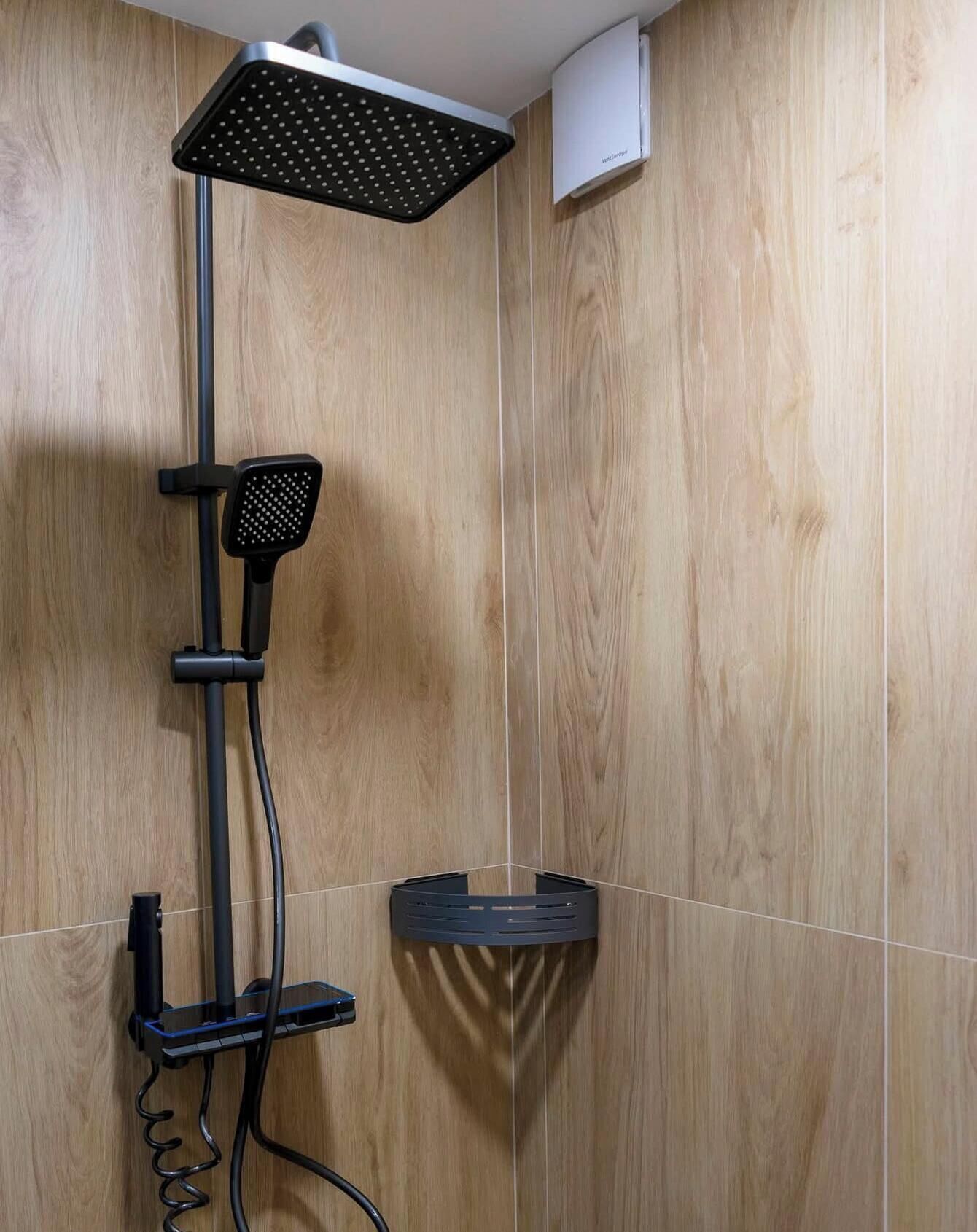 A shower with a handheld showerhead, a wall-mounted control panel, and a corner sink with a black soap dish.