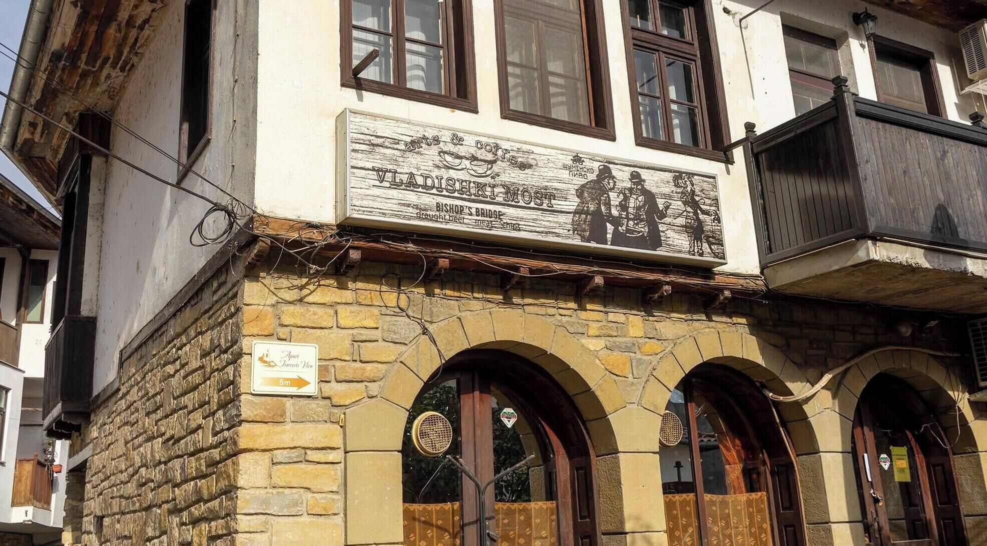 A building with a signboard advertising 'Vladishki Most' and 'Bishop's Bridge' with a view of a bridge.
