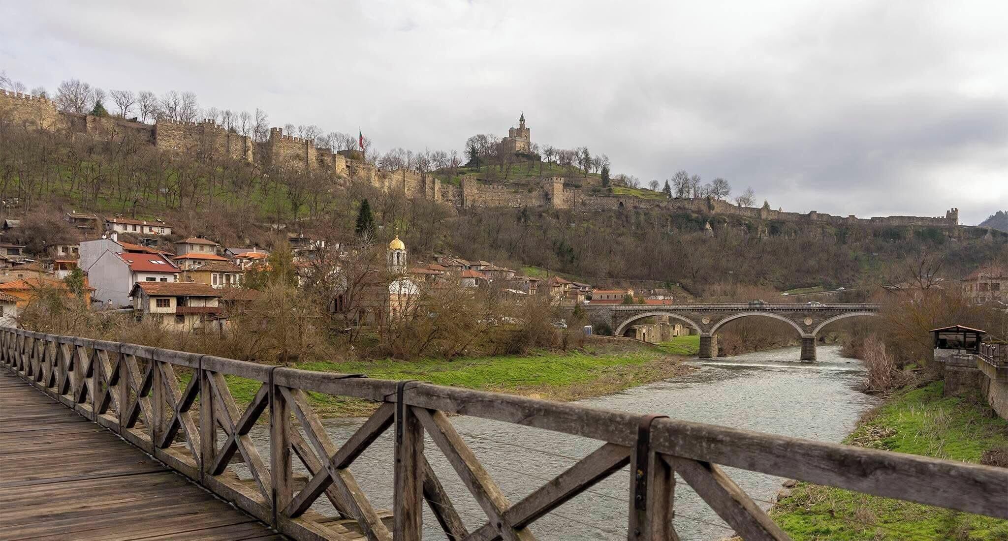 Eine Holzbrücke über einem Fluss, im Hintergrund eine Stadt und ein Schloss.