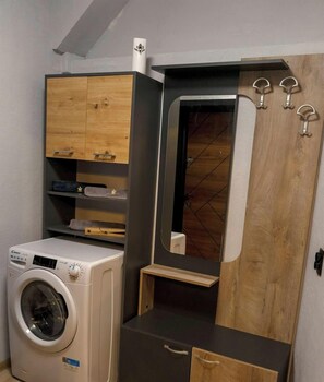 A compact room with a washing machine, wooden cabinets, a mirror, and a small wooden shelf.