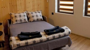 Una cama individual con una manta a cuadros, dos almohadas, una toalla doblada y una mochila sobre la cama. Hay una mesita con un vaso y un recipiente metálico.