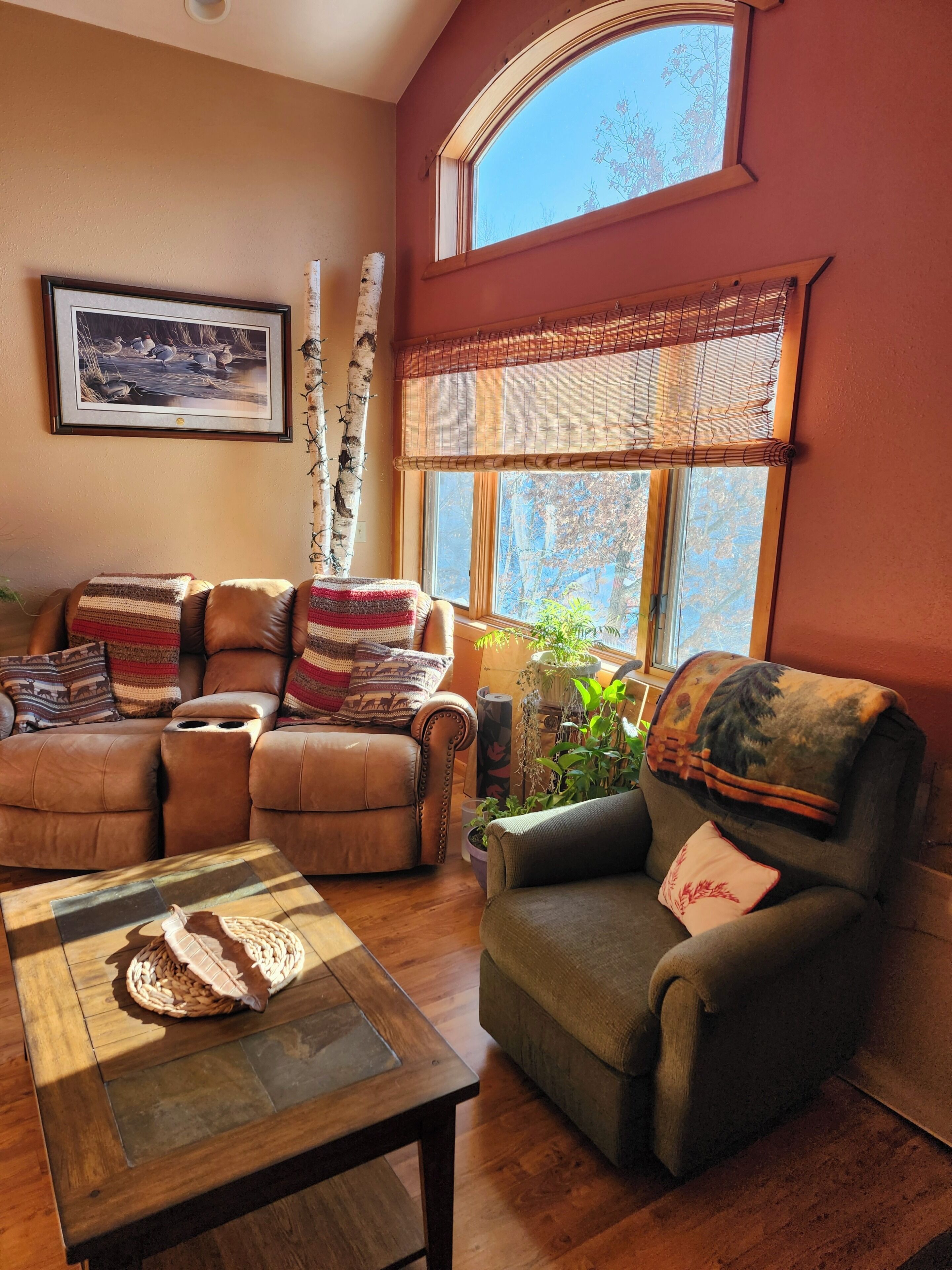 A cozy living room with a leather sofa, a recliner, a coffee table, and a window with a view of trees.