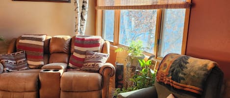 A cozy living room with a leather sofa, a recliner, a coffee table, and a window with a view of trees.