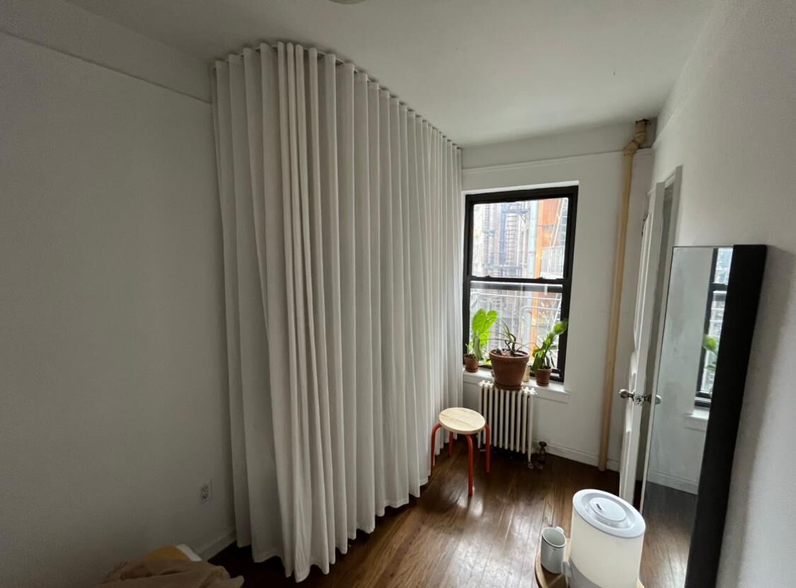 Ein Zimmer mit großem Fenster, Heizkörper, Topfpflanze, kleinem Hocker und einer Tür.