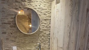 Un bany rústic amb paret de pedra, un tocador de fusta i un mirall rodó.