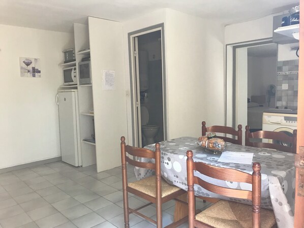 A dining area with a table, chairs, a refrigerator, and a door leading to a kitchen. - Farniente under the olive trees (Santa-Reparata-di-Balagna)