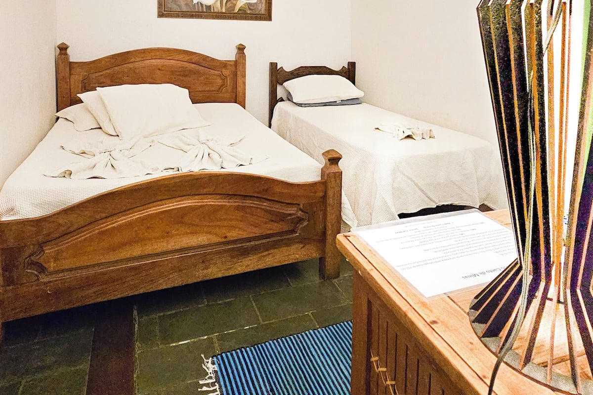 Quarto com duas camas de madeira, um candelabro, um quadro, uma luminária e um tapete azul no chão.