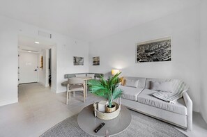 A modern living room with a grey sofa, a coffee table with a plant, a dining area with chairs, and a cityscape painting on the wall. - 1-BD Condo in DTW Miami - City Views (Miami)