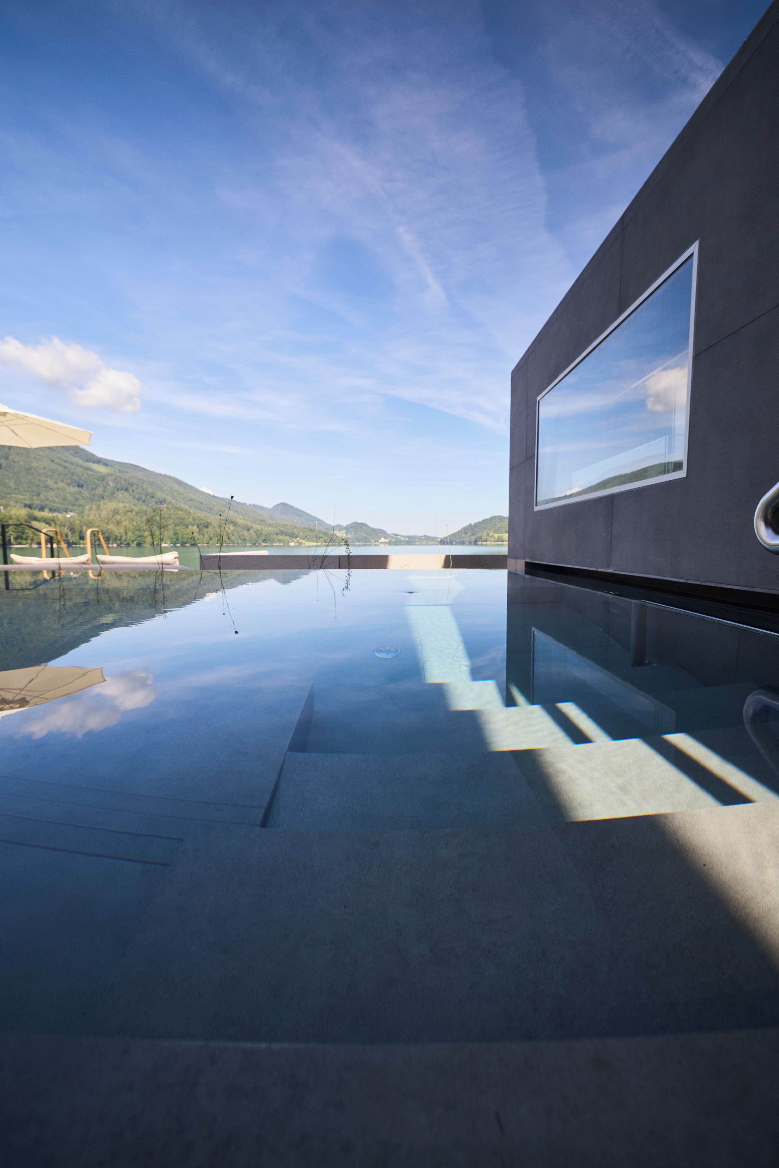 Un edificio moderno con una piscina reflectante, cielo despejado y montañas al fondo.