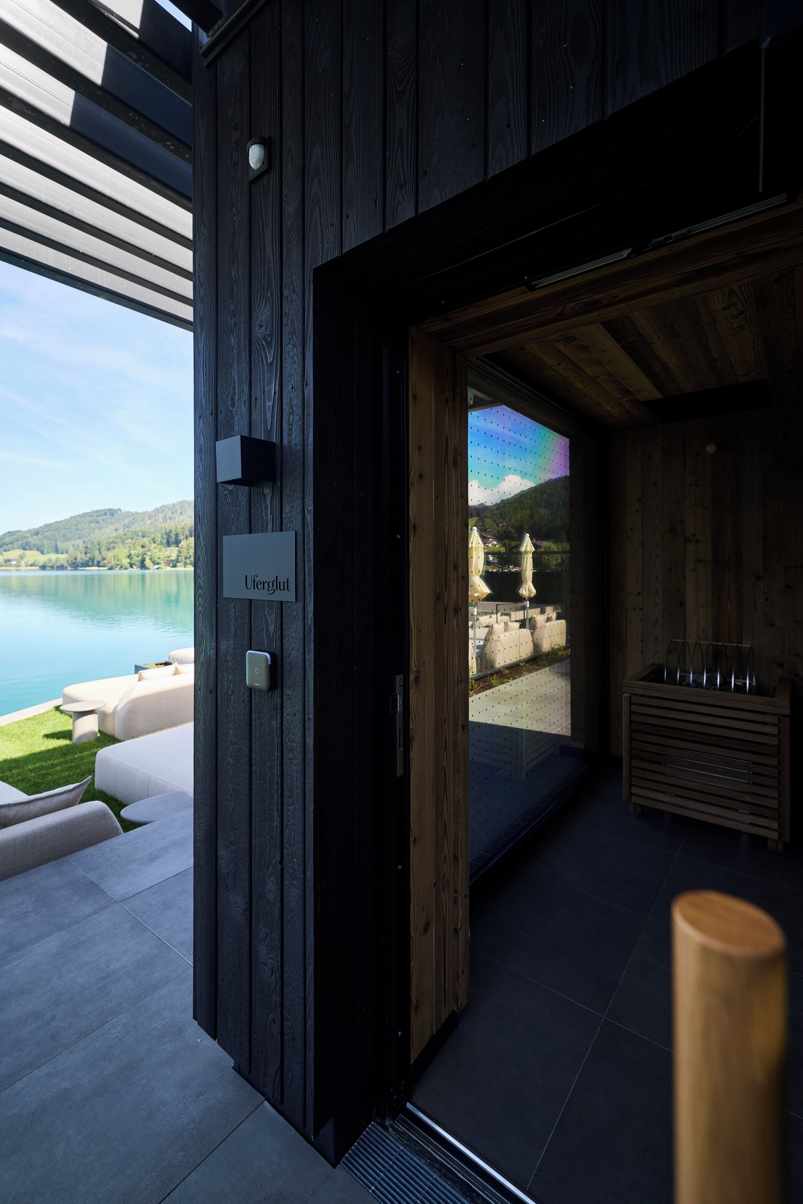 Un edificio moderno con fachada de madera, un cartel que dice 'Uferglut' y vistas a un lago y montañas a través de un vidrio.
