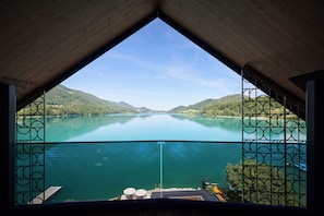 A view from a covered balcony overlooking a calm lake with surrounding hills, a dock with chairs and umbrellas, and a railing with a geometric pattern. - Vogl's Nest (Fuschl am See)