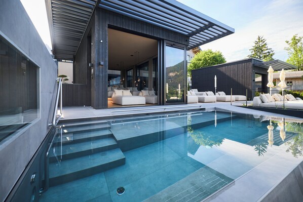 A modern outdoor pool area with a glass-enclosed lounge and a view of the mountains. - Vogl's Nest (Fuschl am See)