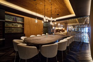 Zona pranzo moderna con un grande tavolo in legno, diverse sedie, lampade a sospensione e un pannello decorativo.