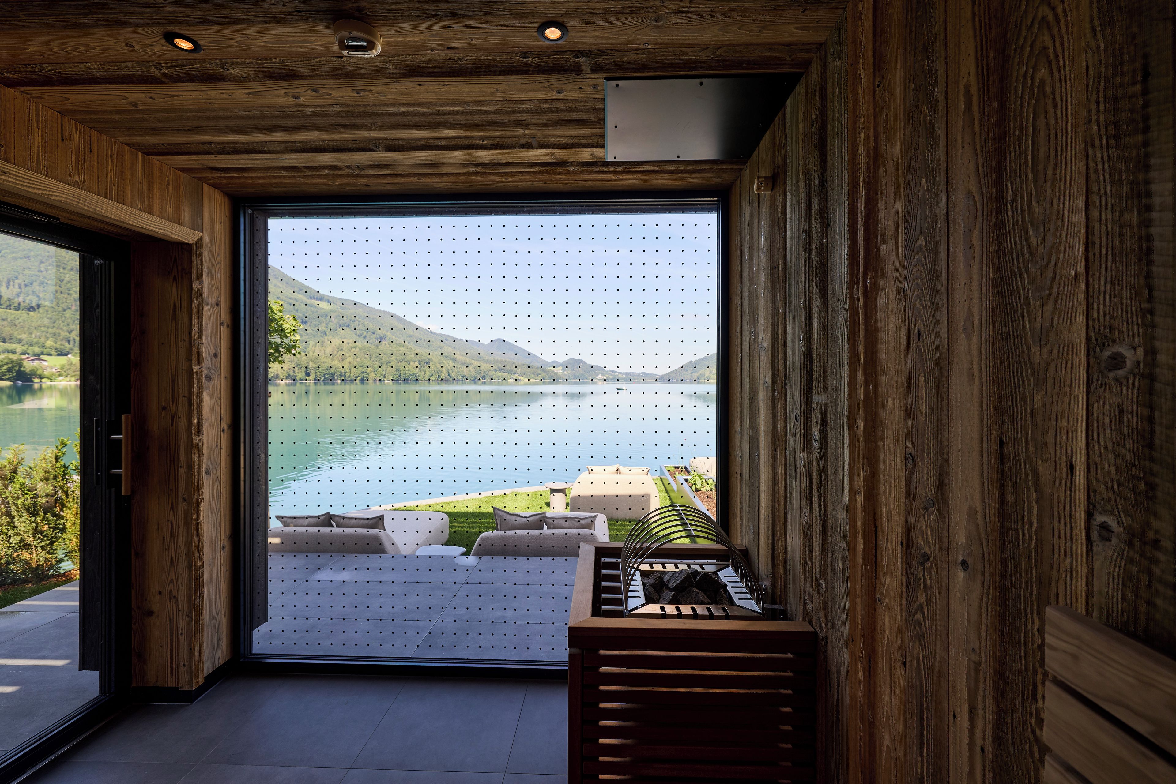 A room with a large window overlooking a lake, a wooden ceiling, and a seating area with cushions.