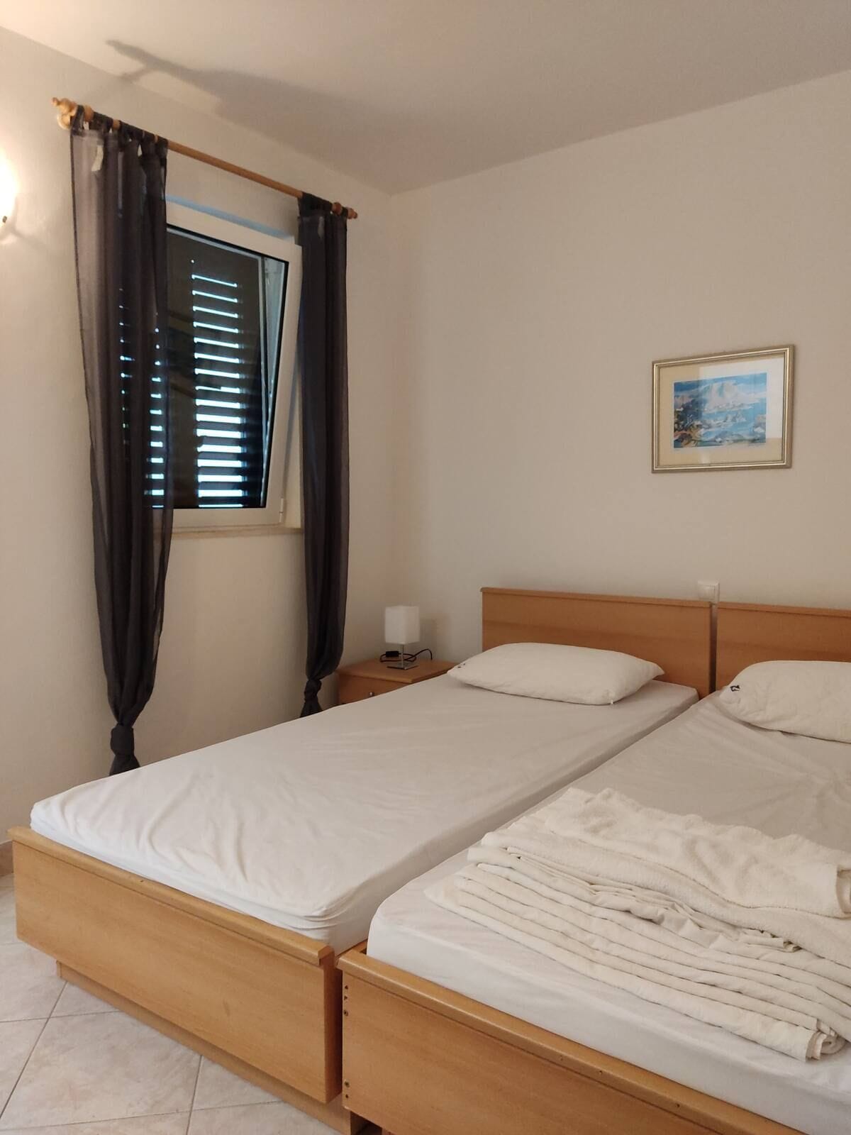 A double bed with a wooden headboard, a nightstand with a lamp, a window with curtains, and a framed picture on the wall.