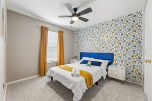 Een slaapkamer met een bed, een nachtkastje, een plafondventilator, een raam met gordijnen en een wand met een patroon.