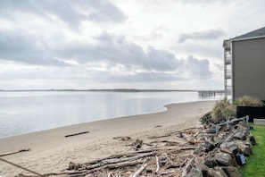 A beach with a wooden pier in the distance, a building with a balcony, and a rocky shoreline. - Dock of the Bay Condo, Fireplace & Bay Views (Lincoln City)