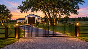 A gated property with a gravel driveway leading to a building with a large tree in front.