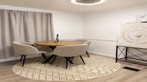 Ein Zimmer mit einem runden Tisch, vier Stühlen, einem kleinen Beistelltisch, einer Wanddekoration und einem Fenster mit Vorhängen.