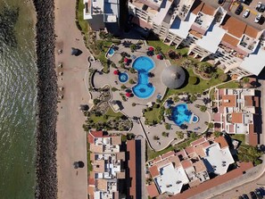 Luftaufnahme eines Küstenhotels mit mehreren Swimmingpools, einem Strand und einem Parkplatz.