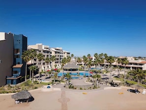Ein Resort mit mehreren Gebäuden, einem zentralen Poolbereich, Palmen und einem Strand mit Liegestühlen.