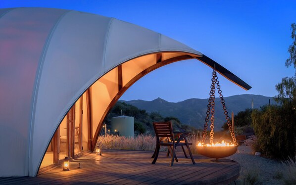 Une structure moderne en forme de tente, dotée d’une terrasse en bois, d’un foyer suspendu et offrant une vue sur les montagnes en arrière-plan.