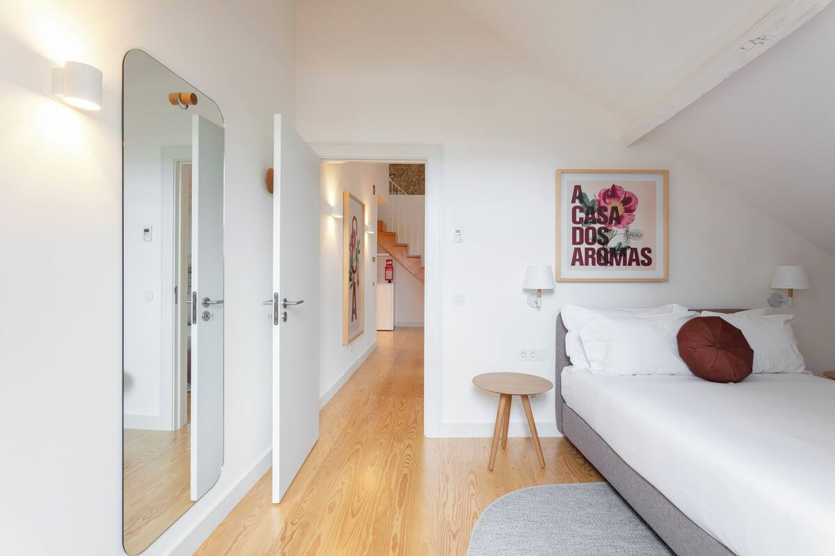 A bedroom with a bed, bedside table, and a mirror. There is a hallway leading to a staircase.