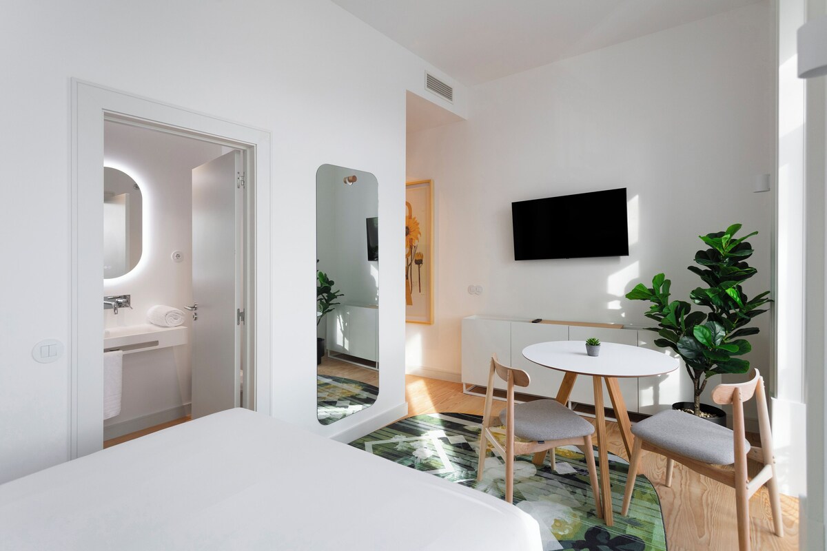 Een hotelkamer met een bed, een klein rond tafeltje met stoelen, een aan de muur bevestigde televisie, een kamerplant en een spiegel.