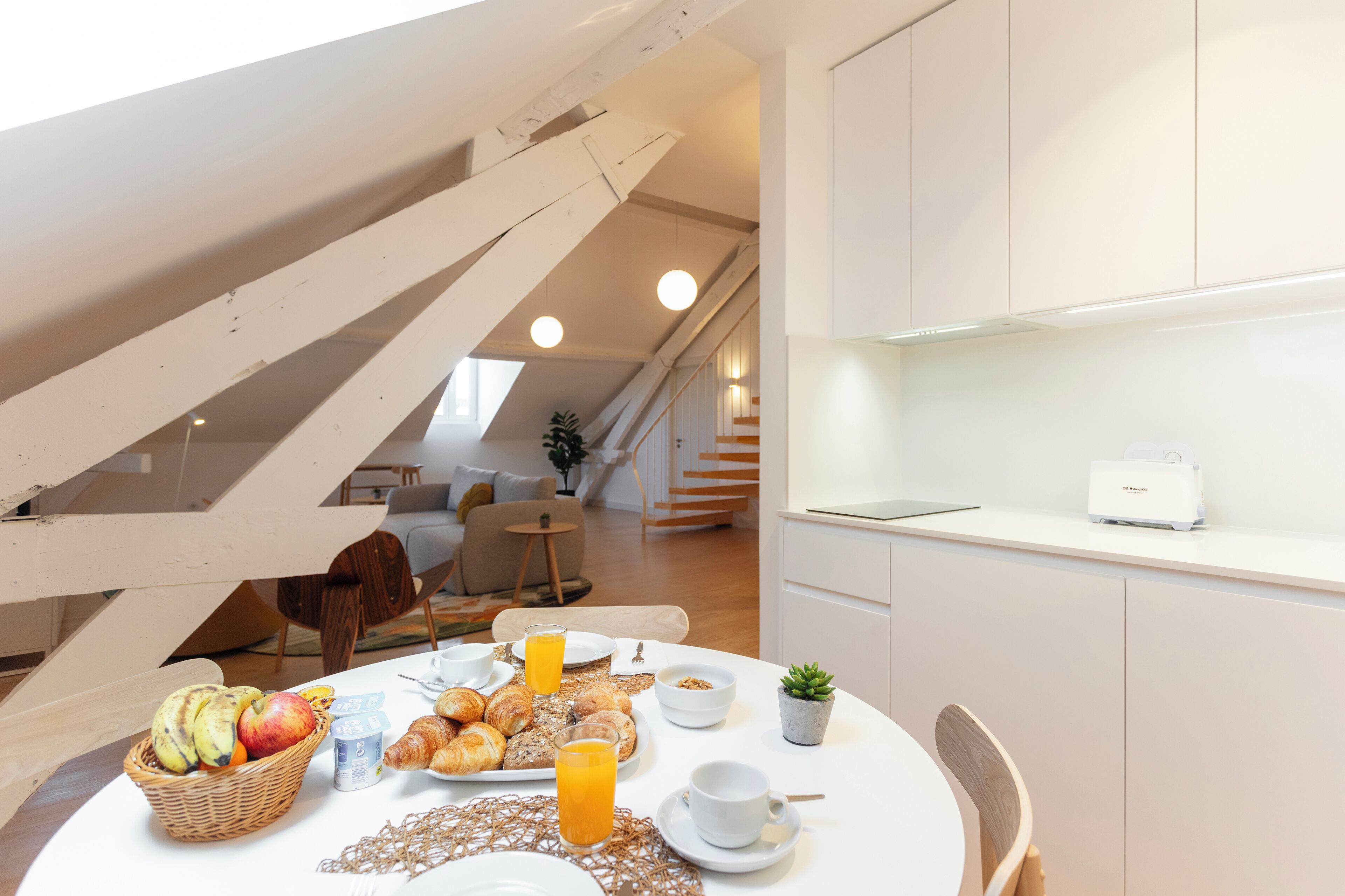 Un desayuno con croissants, fruta y jugo sobre una mesa en una cocina moderna con armarios blancos y una escalera.