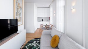 Una sala de estar moderna con un televisor de pantalla plana, un sofá gris con un cojín de color mostaza, un comedor con una mesa redonda y sillas, y una cocina con armarios blancos.