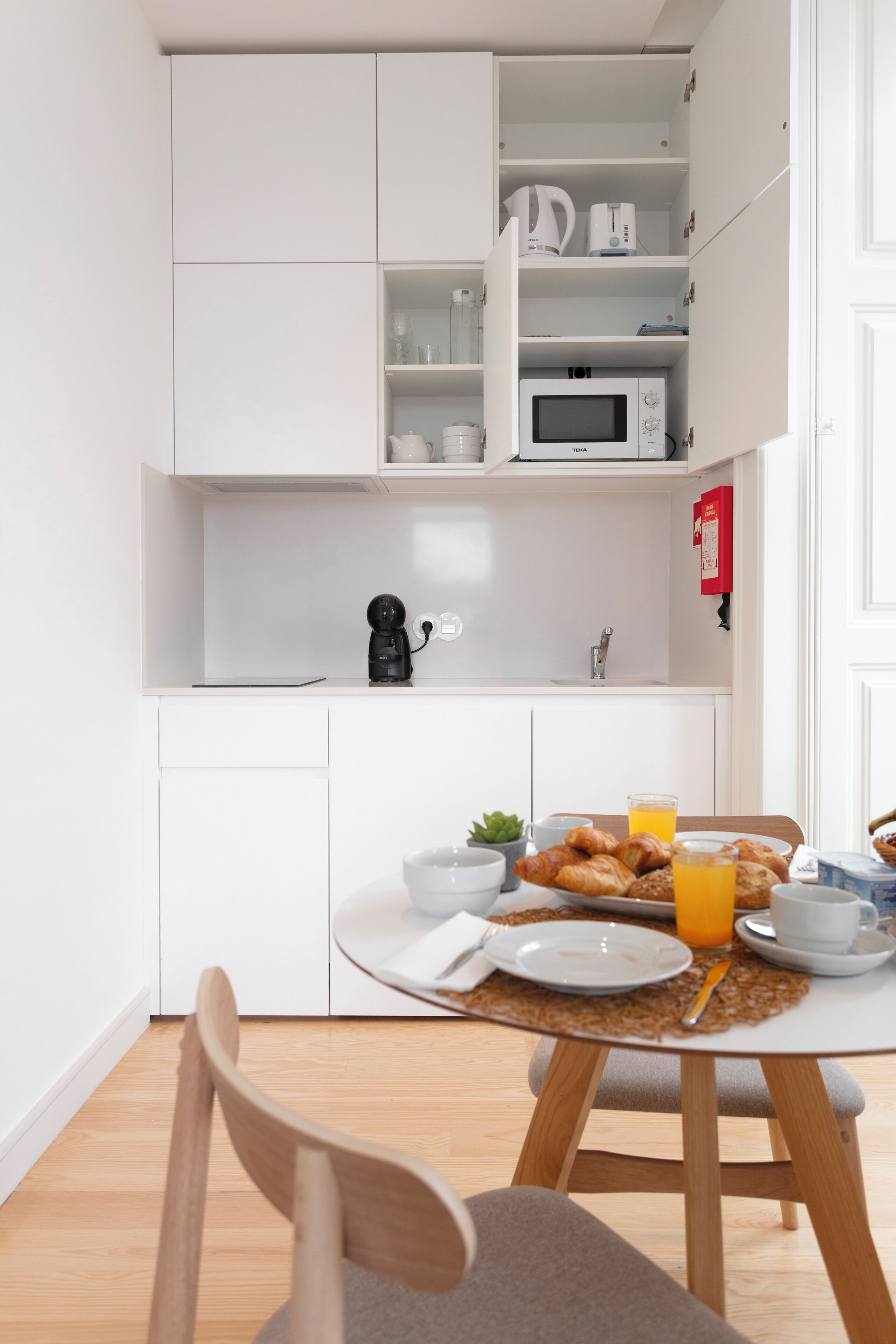 Une cuisine compacte avec des meubles blancs, un four à micro-ondes et une machine à café. Une table est dressée avec des assiettes, une tasse et une bouteille de jus d’orange.