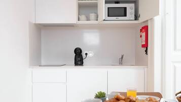 Une cuisine compacte avec des meubles blancs, un four à micro-ondes et une machine à café. Une table est dressée avec des assiettes, une tasse et une bouteille de jus d’orange.