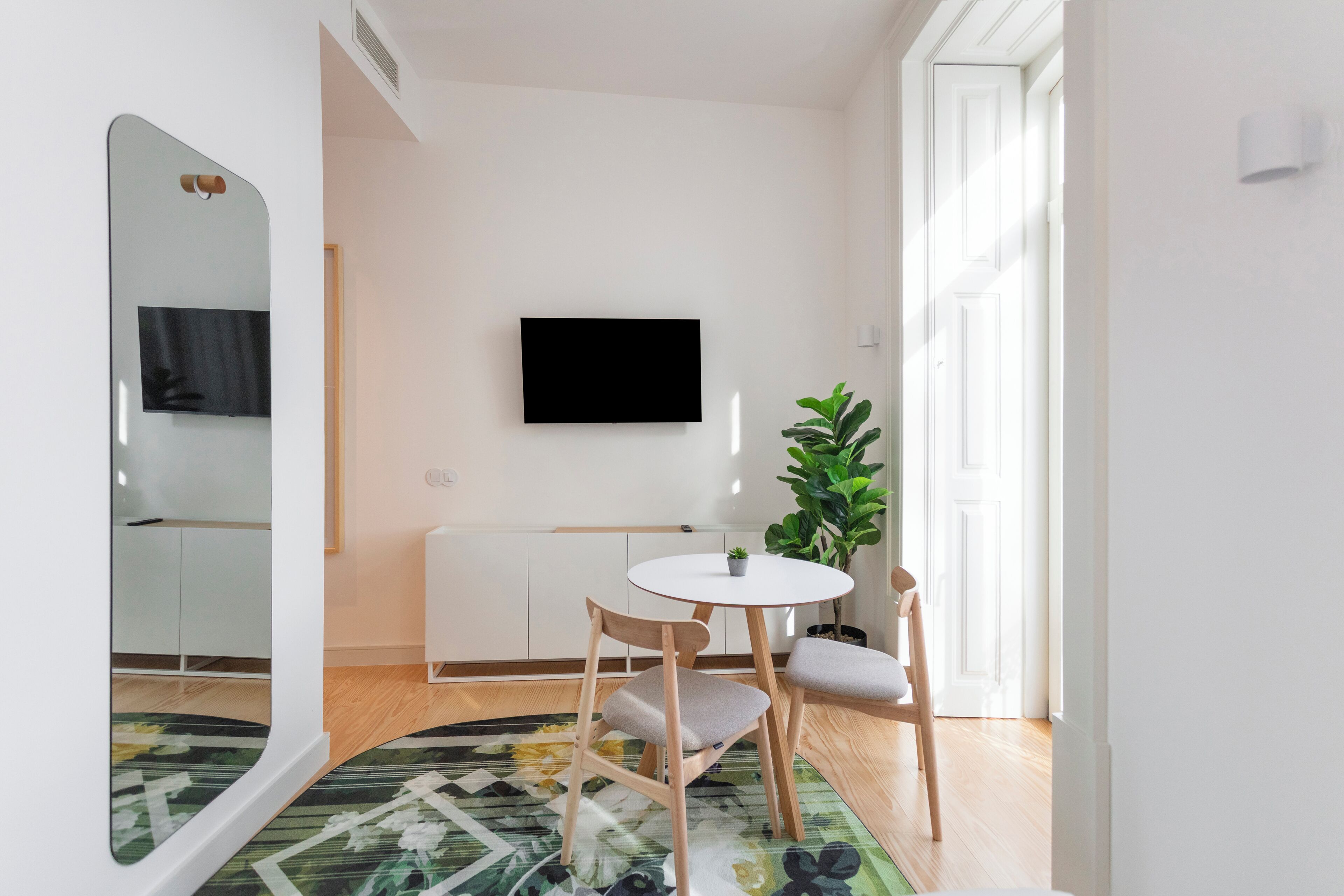 A modern living room with a flat-screen TV, a round table, two chairs, a small plant, and a large mirror.